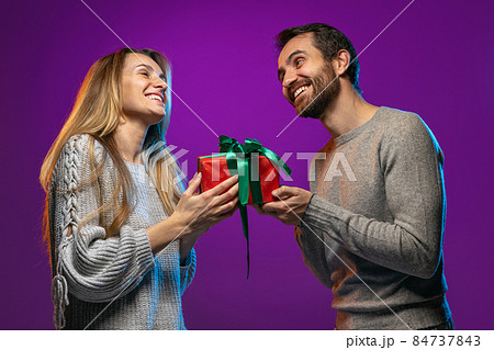Cropped portrait of happy young couple, man giving to woman his holiday present isolated over purple background in neon lights Cropped portrait of happy young couple, man giving to woman his holiday present isolated over purple background in neon lights 84737843
