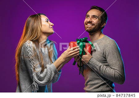 Cropped portrait of happy young couple, woman giving to man his holiday present isolated over purple background in neon lights 84737845