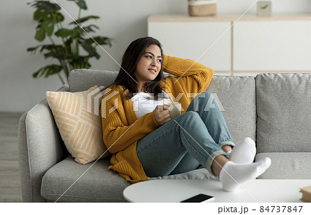Peaceful young arab woman relaxing on sofa with cup of coffee, looking aside and dreaming, free space Peaceful young arab woman relaxing on sofa with cup of coffee, looking aside and dreaming, free space 84737847