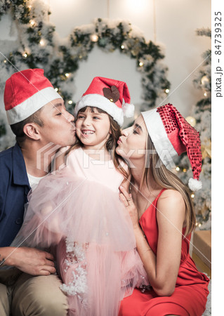 Christmas studio family shoot in red Christmas hats. New Year photo of happy mixed family with cute daughter. Mother and father kiss their daughter Christmas studio family shoot in red Christmas hats. New Year photo of happy mixed family with cute daughter. Mother and father kiss their daughter 84739325