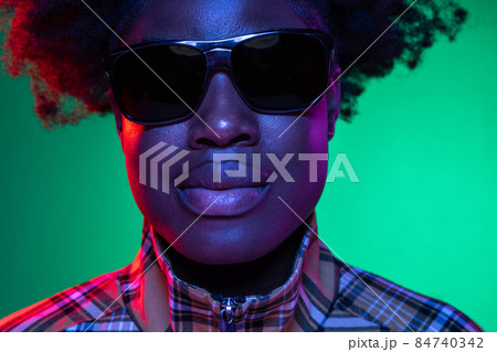 Close-up face of dark skinned young girl in sunglasses isolated on dark green studio background in purple neon light. 84740342