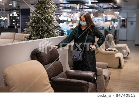 Female customer in mask holding hand on waist and inspecting leather armchair while buying furniture in mall Female customer in mask holding hand on waist and inspecting leather armchair while buying furniture in mall 84740915