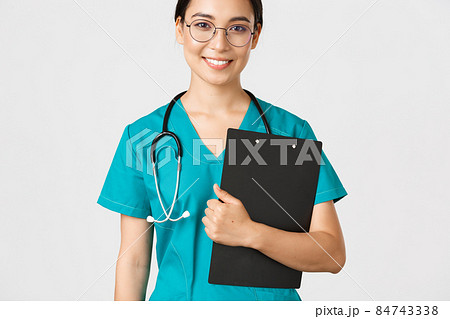 Covid-19, coronavirus disease, healthcare workers concept. Smiling confident asian female nurse, physician in glasses and scrubs holding clipboard, making examination, white background Covid-19, coronavirus disease, healthcare workers concept. Smiling confident asian female nurse, physician in glasses and scrubs holding clipboard, making examination, white background 84743338