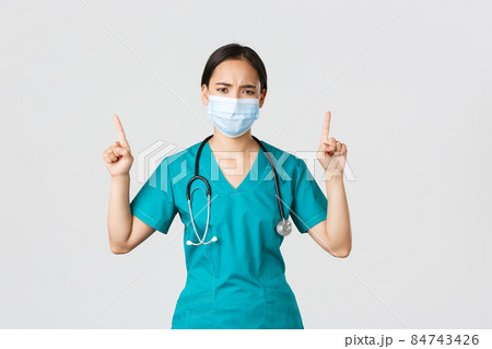 Covid-19, coronavirus disease, healthcare workers concept. Gloomy disappointed asian nurse, physician in medical mask and scrubs, pointing fingers up and frowning perplexed, look frustrated 84743426