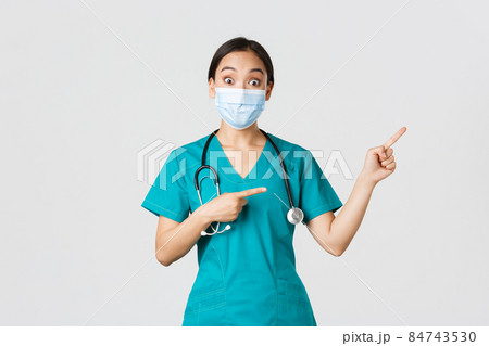 Covid-19, coronavirus disease, healthcare workers concept. Enthusiastic happy asian female doctor, physician or nurse in medical mask and scrubs, pointing upper right corner, show awesome promo 84743530