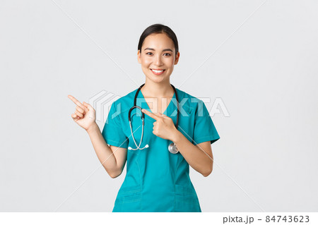 Covid-19, healthcare workers, pandemic concept. Smiling pretty asian doctor, nurse in scrubs and stethoscope, pointing fingers upper left corner, showing online clinic banner, white background Covid-19, healthcare workers, pandemic concept. Smiling pretty asian doctor, nurse in scrubs and stethoscope, pointing fingers upper left corner, showing online clinic banner, white background 84743623