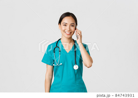 Covid-19, healthcare workers and preventing virus concept. Smiling happy asian female doctor, young intern in scrubs talking on phone, looking at camera hopeful, white background 84743931