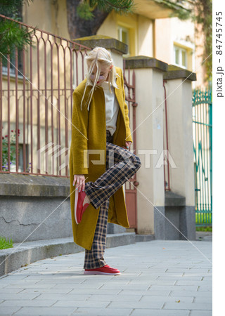 Fashionable beautiful young woman with blond hair in a stylish long coat, checkered pants, red shoes and glasses poses in the city streets. 84745745