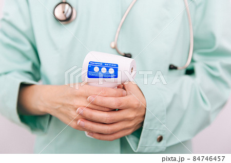 Temperature measurement gun in doctor hands. Close-up shot of doctor wearing protective surgical mask ready to use infrared isometric thermometer gun to check body temperature for virus symptoms. Temperature measurement gun in doctor hands. Close-up shot of doctor wearing protective surgical mask ready to use infrared isometric thermometer gun to check body temperature for virus symptoms. 84746457