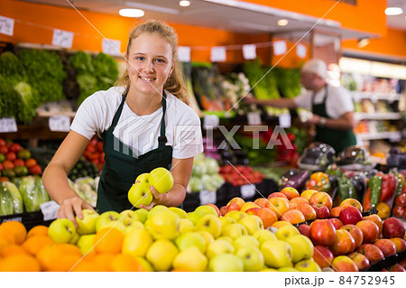 Fifteen-year-old girl who works part-time as a trainee saleswoman puts apples on the counter Fifteen-year-old girl who works part-time as a trainee saleswoman puts apples on the counter 84752945