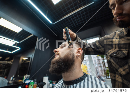 Barber works with a beard clipper. Hipster client getting haircut. Barber works with a beard clipper. Hipster client getting haircut. 84764976