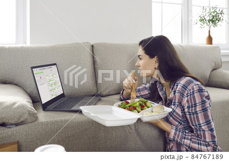 Serious girl using timetable organizer app on modern laptop computer on her lunch break. Hungry woman with busy schedule eating fresh meal from plastic food container ordered in fast takeaway delivery Serious girl using timetable organizer app on modern laptop computer on her lunch break. Hungry woman with busy schedule eating fresh meal from plastic food container ordered in fast takeaway delivery 84767189