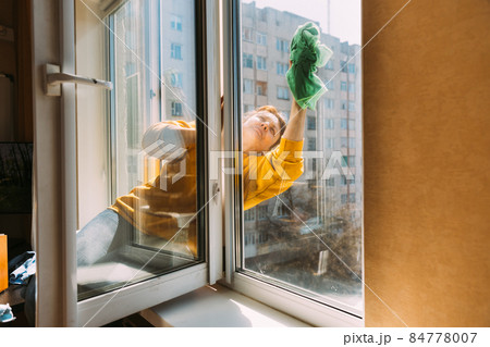 Caucasian Woman Of Fifty In A Yellow Sweater And Jeans Washes A Dusty Window In Apartment. A 50 Year Old Woman Cleans Windows From Stains Using Rag. Elderly Woman Is Cleaning House, Doing Household Caucasian Woman Of Fifty In A Yellow Sweater And Jeans Washes A Dusty Window In Apartment. A 50 Year Old Woman Cleans Windows From Stains Using Rag. Elderly Woman Is Cleaning House, Doing Household 84778007