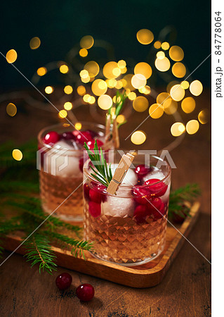 Alcoholic cocktail or non-alcoholic cocktail with vodka and cranberries with ice with fir branches and glowing garland for christmas on wooden background Alcoholic cocktail or non-alcoholic cocktail with vodka and cranberries with ice with fir branches and glowing garland for christmas on wooden background 84778064