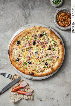 Tasty pizza with sausage, mozzarella, marinated mushrooms, black olives, green onion on marble round tray. Garnished on grey stone backdrop with pizza ingredients Tasty pizza with sausage, mozzarella, marinated mushrooms, black olives, green onion on marble round tray. Garnished on grey stone backdrop with pizza ingredients 84779526
