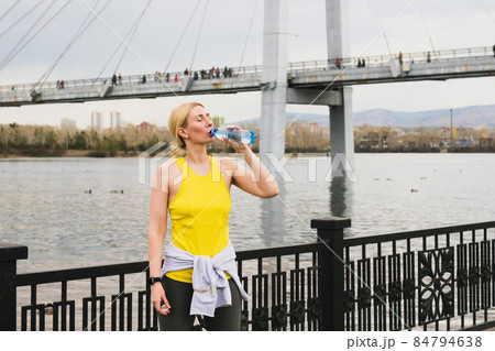 A blonde middle-aged woman in bright sportswear athlete drinks water while resting during a sports training the street A blonde middle-aged woman in bright sportswear athlete drinks water while resting during a sports training the street 84794638