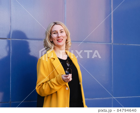 Young woman with blond hair in a bright yellow coat on a blue background on a city street. Urban style of modern woman Young woman with blond hair in a bright yellow coat on a blue background on a city street. Urban style of modern woman 84794640