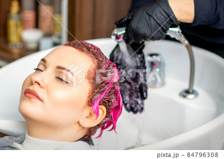 Washing dyed female hair. Professional hairdresser washes pink color paint off of a customer. Young caucasian woman having her hair washed in a beauty salon. 84796308