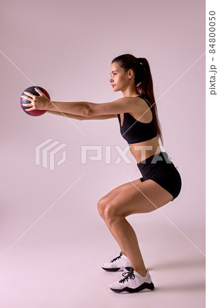 Side view portrait of Slim caucasian cross fit woman with fitness ball doing squats, isolated Side view portrait of Slim caucasian cross fit woman with fitness ball doing squats, isolated 84800050