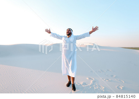 Happy Muslim man looks through banknotes and beside himself with joy, standing in center of sandy desert on warm, hot day. Happy Muslim man looks through banknotes and beside himself with joy, standing in center of sandy desert on warm, hot day. 84815140