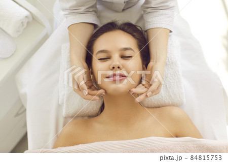 Happy young woman relaxing on massage bed or table at spa center, getting face massage, and enjoying rejuvenating facial treatment for bright complexion, even tone and clear fresh perfect glowing skin 84815753