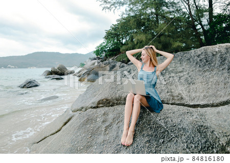 Young blonde woman working with laptop on sea shore and sitting on stone. Young blonde woman working with laptop on sea shore and sitting on stone. 84816180