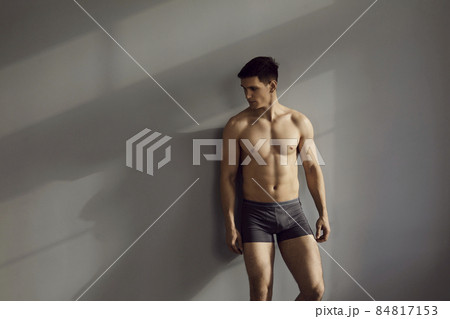 Sexy portrait of muscular shirtless male model in underwear posing in sensual pose against gray wall background. Caucasian brunette man in black cotton underpants looks away while standing in sunlight 84817153
