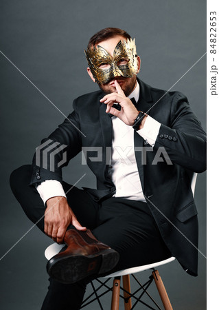 Man in business suit sits on chair wearing carnival mask on his face. Studio portrait on gray Man in business suit sits on chair wearing carnival mask on his face. Studio portrait on gray 84822653
