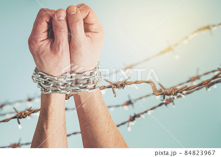Hands tied with metal chain raised against barbed wire fence 84823967