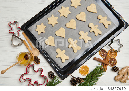 Top view of laid out gingerbread cookies on pastry rack. Cookies cutter, honey, pine scones and branches, dried slices of orange on wooden surface. Cooking process. Christmas preparations. 25 December 84827050