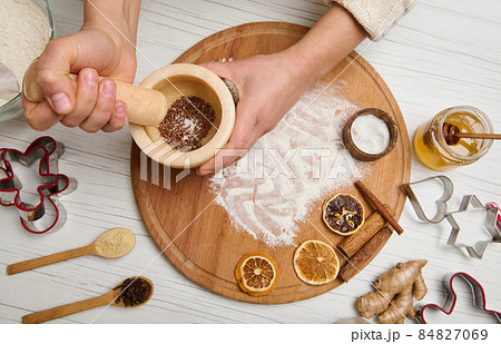 Woman's hands grinding ingredients in a marble mortar standing on a wooden board with sprinkled flour and dried orange slices, cinnamon sticks lying on it, cutting molds on a wooden table 84827069
