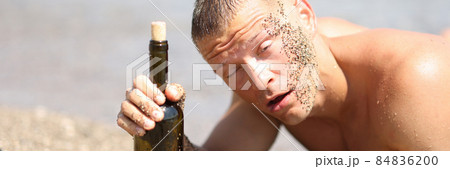 Drunk young man in sand lying on beach with bottle of wine in his hands Drunk young man in sand lying on beach with bottle of wine in his hands 84836200