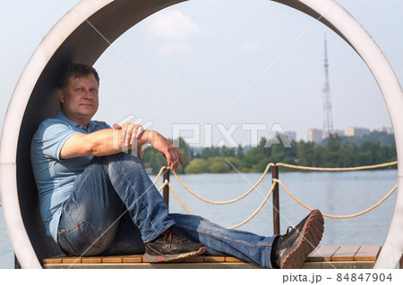 An adult man sits on a creative bench in the form of a circle and looks at the calm surface of the river An adult man sits on a creative bench in the form of a circle and looks at the calm surface of the river 84847904