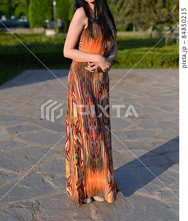 beautiful brunette model with long hair is having a photo shooting in the park. the woman is wearing an orange long summer dress beautiful brunette model with long hair is having a photo shooting in the park. the woman is wearing an orange long summer dress 84850215