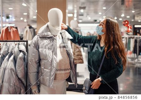 Young lady in face mask chooses warm clothes in department store, standing near mannequin dressed in puffy winter jacket and sweater, buying new seasonal outfit. Shopping during covid19 pandemic 84856856