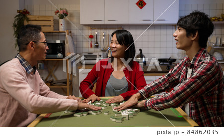 happy mother chatting with father and son while they are playing mahjong together in the evening during spring festival. text on cabinet translation: luck 84862035