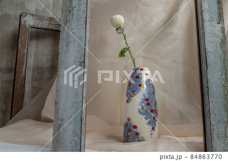 White flower in Blue and White, Red, Yellow handmade ceramic vase on Blush textured table cloth. White flower in Blue and White, Red, Yellow handmade ceramic vase on Blush textured table cloth. 84863770