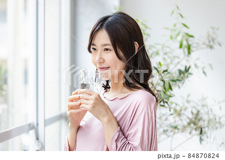 水の入ったグラスを持って窓際に立つルームウェアの女性 84870824