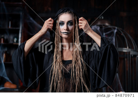 Halloween concept. Witch portrait close up with dreadlocks looking camera dressed black hood standing dark room with cage on background 84870971