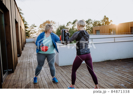 Overweight woman training boxing with personal trainer outdoors on terrace. 84871338