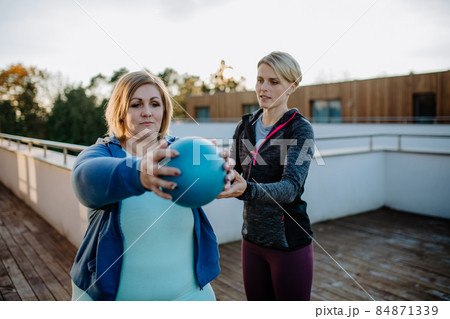 Overweight woman holding ball and exercising with personal trainer in outdoors on gym terrace. 84871339