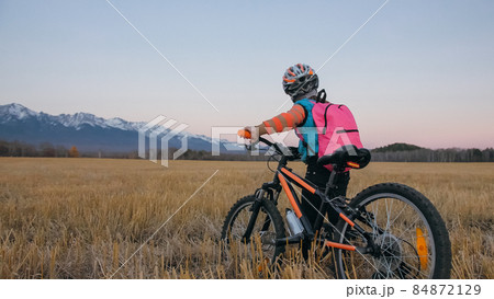 One caucasian children walk with bike in wheat field. Little girl walking black orange cycle on background of beautiful snowy mountains. Biker stand with backpack and helmet. 84872129