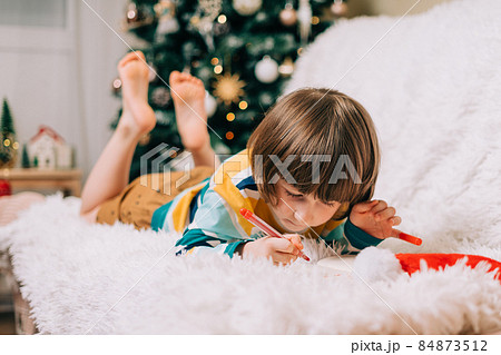 Smiling Kid boy lying on couch and writing the letter to dear Santa at home near Christmas tree. Child wish list. Dreams of a Christmas gifts. Merry Christmas and Happy new year. 84873512