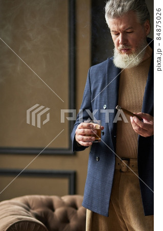 Cropped shot of serious mature man with beard looking at cigar while holding it and glass of whisky, standing in loft interior Cropped shot of serious mature man with beard looking at cigar while holding it and glass of whisky, standing in loft interior 84875026
