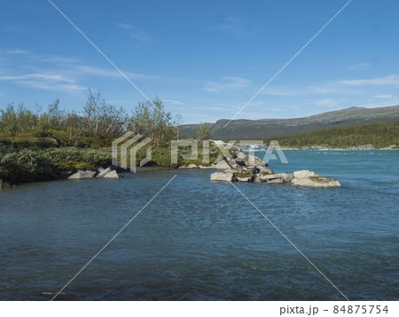 View of turquoise blue water of wild Vuojatadno river. Northern landscape in Swedish Lapland with birch tree forest and green mountains at Padjelantaleden hiking trail. Summer sunny day, blue sky 84875754
