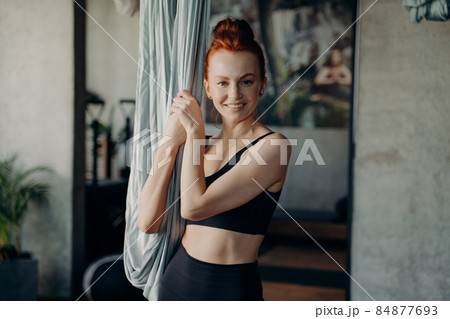 Positive ginger girl standing near hammock smiling on camera waiting for aerial yoga class 84877693