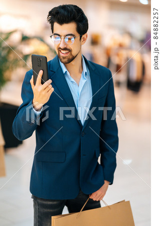 Vertical portrait of handsome bearded young man in fashionable suit and glasses using mobile phone standing in hall of mall, holding shopping bags. 84882257