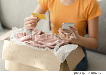 Modern home shopping. Young unrecognizable woman with credit card and phone, unpacking cardboard box with clothes Modern home shopping. Young unrecognizable woman with credit card and phone, unpacking cardboard box with clothes 84882876