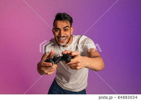 Handsome young Arab man using joystick, playing online video game in neon light Handsome young Arab man using joystick, playing online video game in neon light 84883034
