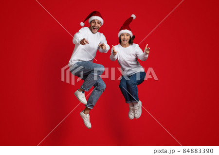 Overjoyed Man And Woman In Santa Hats Jumping And Pointing At Camera Overjoyed Man And Woman In Santa Hats Jumping And Pointing At Camera 84883390